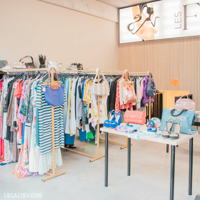 Intérieur de la boutique Les Fééries by Val-Lou, avec des portants de vêtements et une table exposant des accessoires de mode, sacs et chaussures, dans un espace lumineux et soigné.