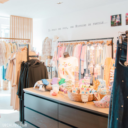 Intérieur du magasin - Vue d'ensemble : Un espace lumineux et moderne, avec des portants de vêtements aux tons pastel et neutres. Sur un comptoir en bois noir, plusieurs accessoires de mode, tels que des chouchous, des bracelets et des écharpes, sont joliment disposés dans des paniers en osier. Une inscription sur le mur indique "Il était une fois, une histoire de famille...".