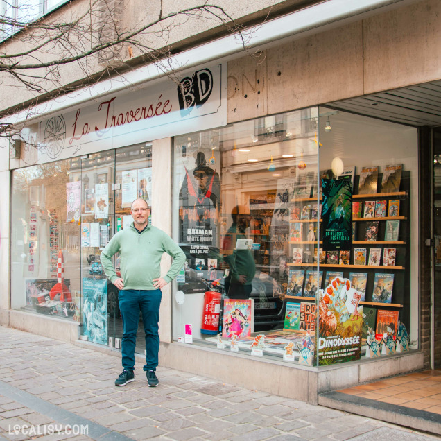 Devanture de la librairie "La Traversée BD", avec un homme en pull vert et pantalon bleu debout devant la vitrine. La vitrine est décorée avec des bandes dessinées et des affiches promotionnelles.