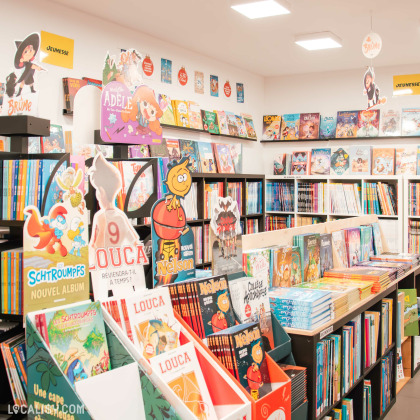 Espace jeunesse de la librairie, avec une décoration colorée et des présentoirs mettant en avant des bandes dessinées pour enfants comme "Schtroumpfs" et "Louca". Les étagères regorgent de BD adaptées aux jeunes lecteurs.