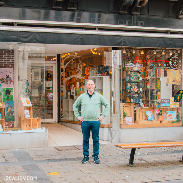 Devanture de la librairie "La Traversée Jeunesse", spécialisée en livres pour enfants et adolescents. Un homme en pull vert et pantalon bleu se tient devant l’entrée. La vitrine est décorée avec des albums jeunesse et des illustrations colorées.