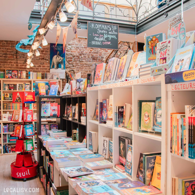 Intérieur de la librairie "La Traversée Jeunesse", mettant en avant un large choix de livres pour jeunes lecteurs. Des étagères blanches et noires sont remplies de romans et albums illustrés. Une pancarte suspendue indique différentes catégories comme "Romans jeunes adultes, livres-jeux, blagues, énigmes".