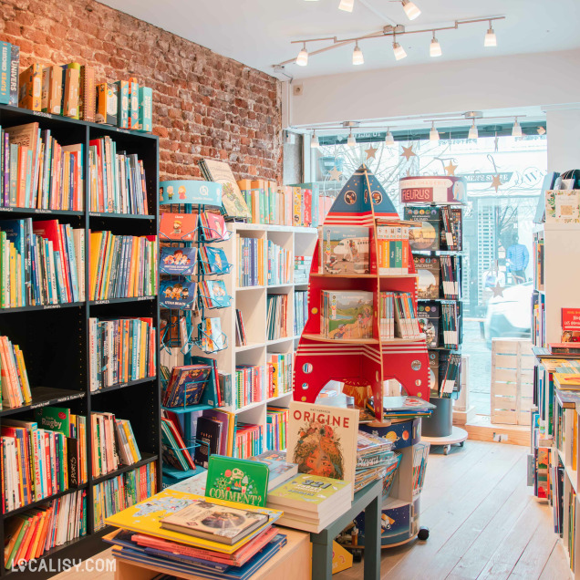 Vue intérieure de la librairie jeunesse avec un décor chaleureux, mur en briques apparentes et sol en bois. Les livres sont disposés sur des étagères et un présentoir en forme de fusée rouge attire l’attention. La lumière naturelle éclaire l’espace depuis la vitrine.