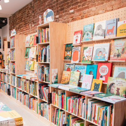 Rayon jeunesse de la librairie avec des étagères en bois remplies d’albums illustrés. Des livres colorés sont exposés en face avant pour attirer les jeunes lecteurs. Le mur en briques apporte un charme authentique à l’espace.