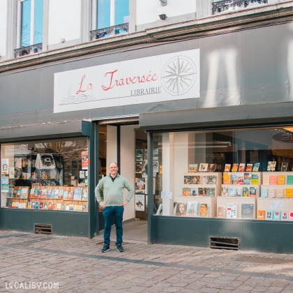 Devanture de la librairie "La Traversée", spécialisée en littérature et essais. Un homme en pull vert et pantalon bleu se tient devant l’entrée. La vitrine met en avant une sélection de livres soigneusement exposés.