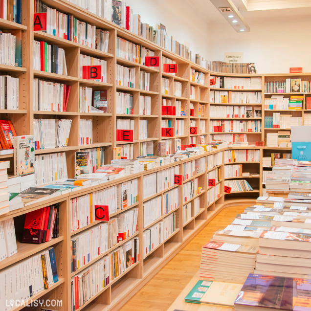 Rayons de la librairie avec une organisation alphabétique des ouvrages sur des étagères en bois clair. De nombreux livres sont disposés en face avant pour attirer l’œil des lecteurs.