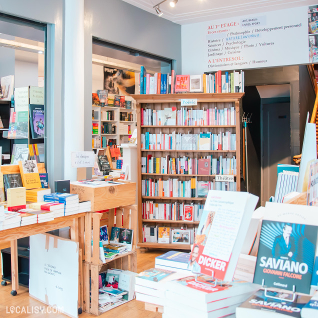 Espace dédié à la poésie et au théâtre dans la librairie, avec des étagères remplies de livres et des tables présentant des sélections thématiques. Un panneau mural indique les différentes sections disponibles à l’étage.