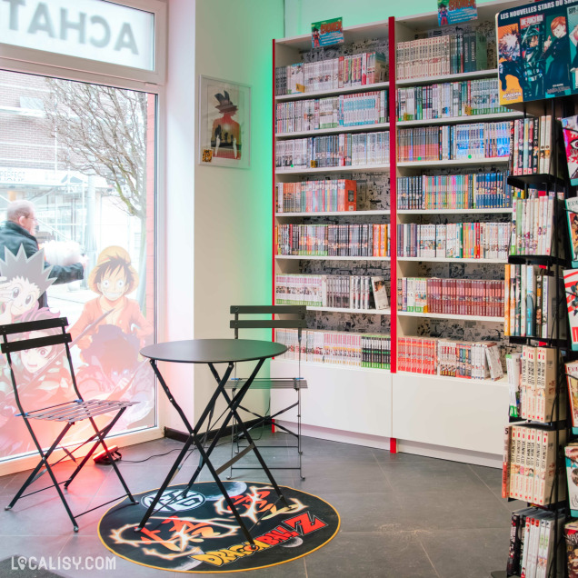 Un coin lecture cosy chez Manga Cash avec une table et deux chaises près d’une vitrine décorée de personnages de mangas populaires. Les étagères en arrière-plan débordent de mangas.